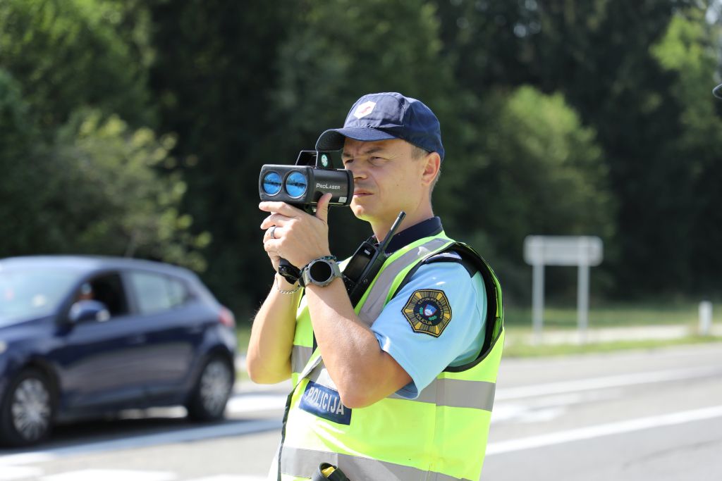 Policist meri hitrost, glede skozi radar, ki ga drži v roki.