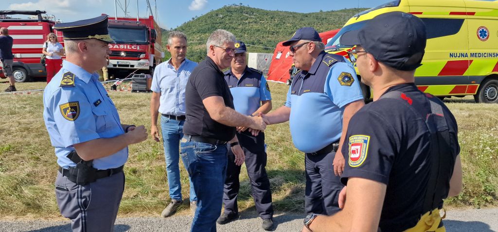 Minister Boštjan Poklukar se rokuje s policisti v uniformah. Stojijo na prostem na travi, zadaj rdeče gasilsko vozilo, v daljavi porasel hrib.