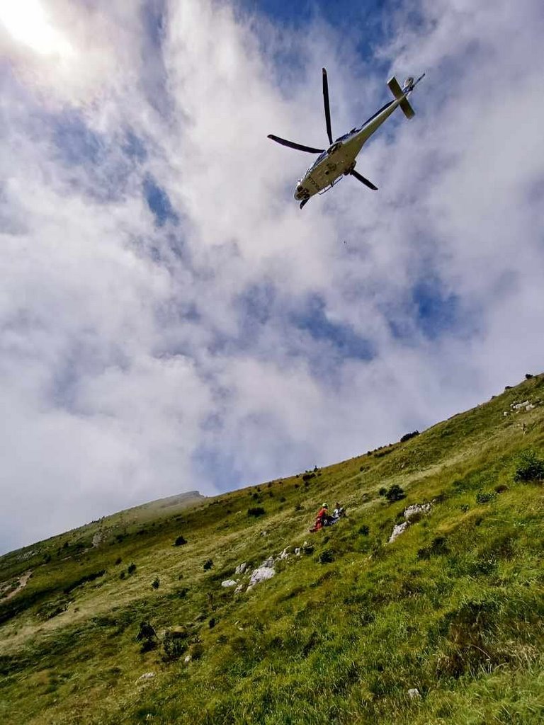Policijski helikopter leti nad strmim travnatim pobočjem, kjer reševalec navezuje poškodovanca