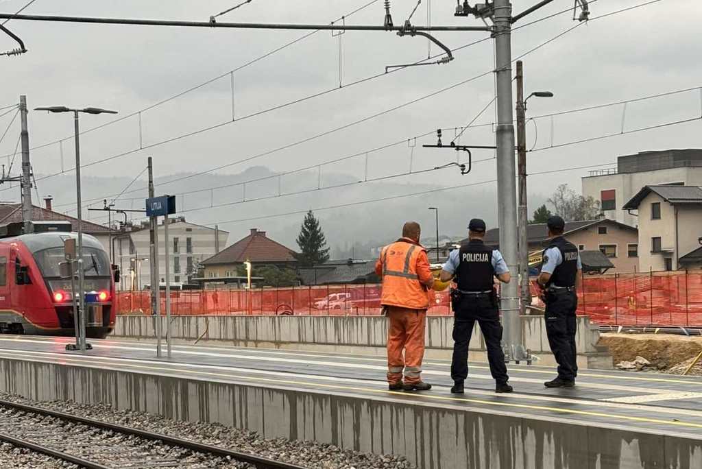 Policista skupaj z delavcem Slovensklih železnic stojita na železniški postaji ob progi, v ozadju se v smeri proti njim približuje rdeč vlak