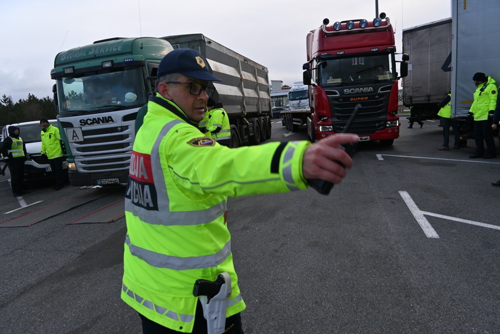 V ospredju policist - vodja nadzora v rumeni vetrovki, v ozadju nekaj parkiranih tovornjakov na avtocestnem počivališču