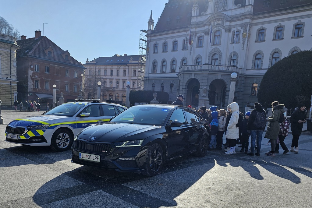 Skupina otrok in odraslih si ogleduje policijski vozili (patruljno in civilno) in se pogovarja s policistom.