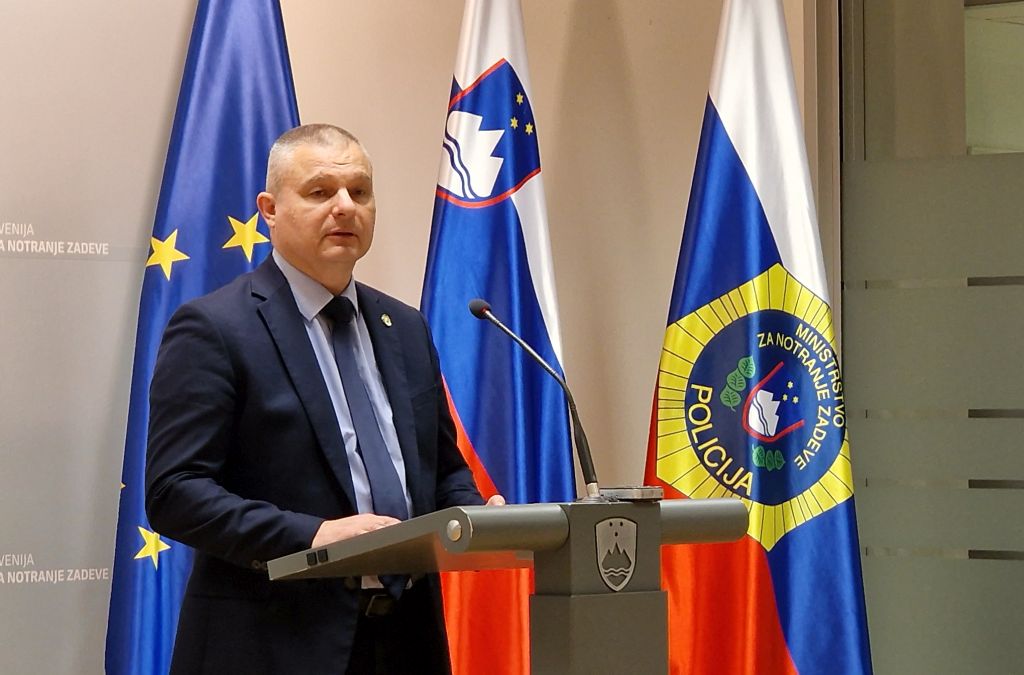 Darko Muženič stoji pred mikrofonom med izjavo v medijskem središču ministrstva, za njim zastave