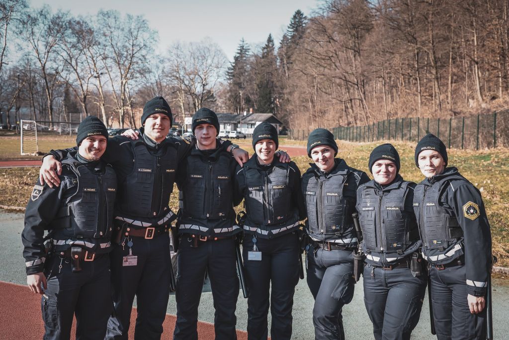 Skupina osmih nasmejanih kandidatov za policiste stoji zunaj pred Policijsko akademijo, v ozadju tekaška proga.