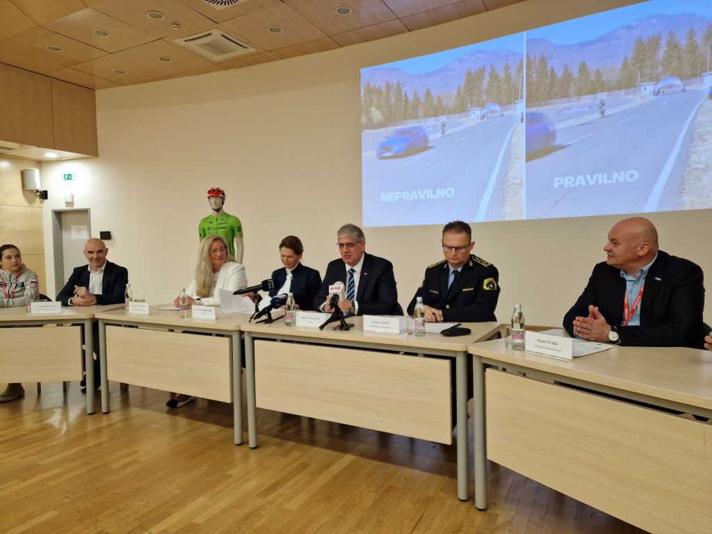 Govorci na novinarski konferenci, sedijo za mizo, za njimi v ozadju se predvaja videoprojekcija.