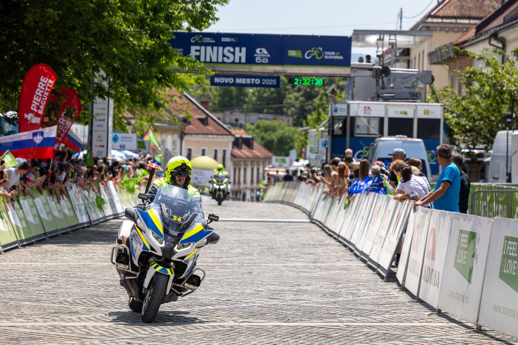 Policist motorist se je pripeljal v cilj zadnje etape, ob progi stojijo navijači, v ozadju nad progo sta obešena napisa Finish in Novo mesto. Foto Gregor Stopar