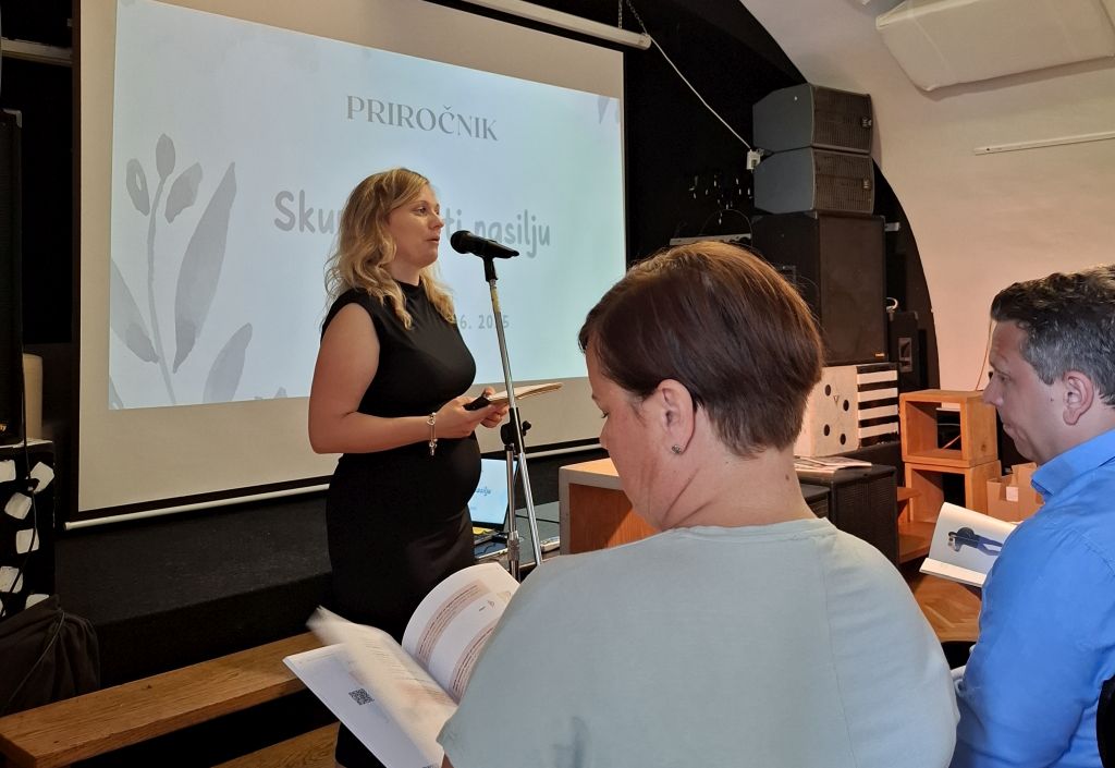Nina Ukmar Mezinec med predstavitvijo priročnika, stoji pred mikrofonom in govori, pred njo so poslušalci.