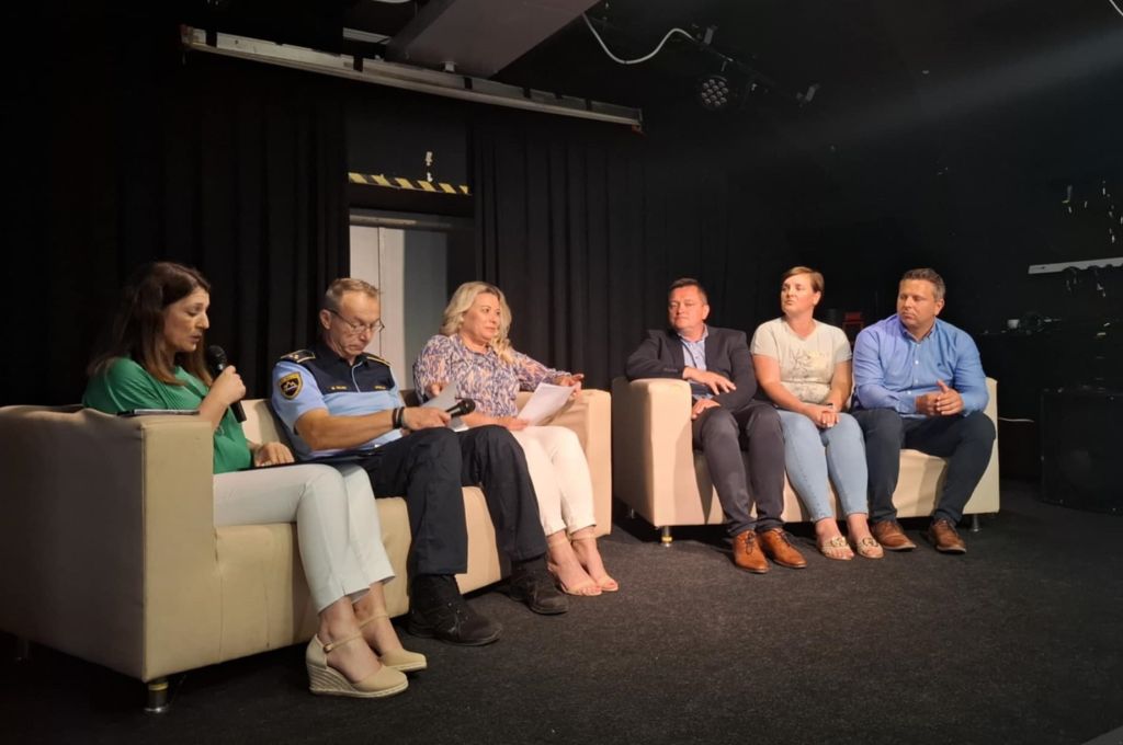 Šest sodelujočih sedi na odru v polkrogu, voditeljica ima v roki mikrofon in nagovarja udeležence.