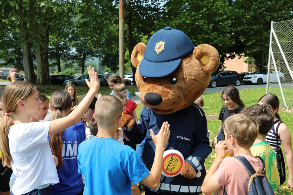 V parku Policijske akademije stoji maskota, medo policist, okrog njega so zbrani nasmejani otroci, z rokami si dajejo "petke".
