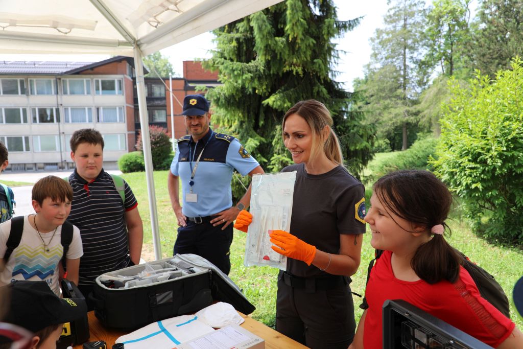 Skupina otrok je zbranih okoli stojnice, kjer jim kriminalistična tehnica kaže svoje pripomočke, poleg nje stoji še en splošni policist.