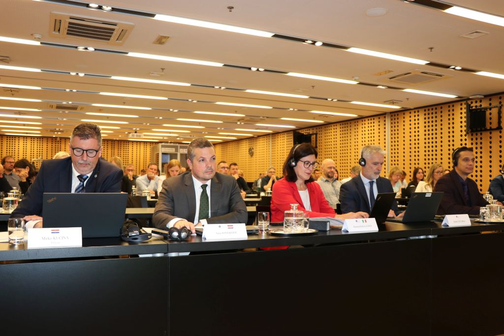 Predstavniki različnih institucij sedijo vsak za svojo mizo v kongresni dvorani in aktivno sodelujejo pri razpravi, pred vsakim je tablica z imenom in priimkom.