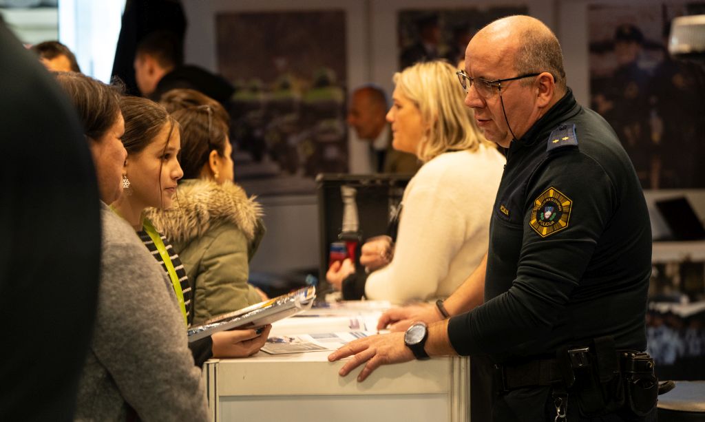 Obiskovalci na razstavnem prostoru stojijo ob pultu in se pogovarjajo s starejšim policistom na drugi strani.
