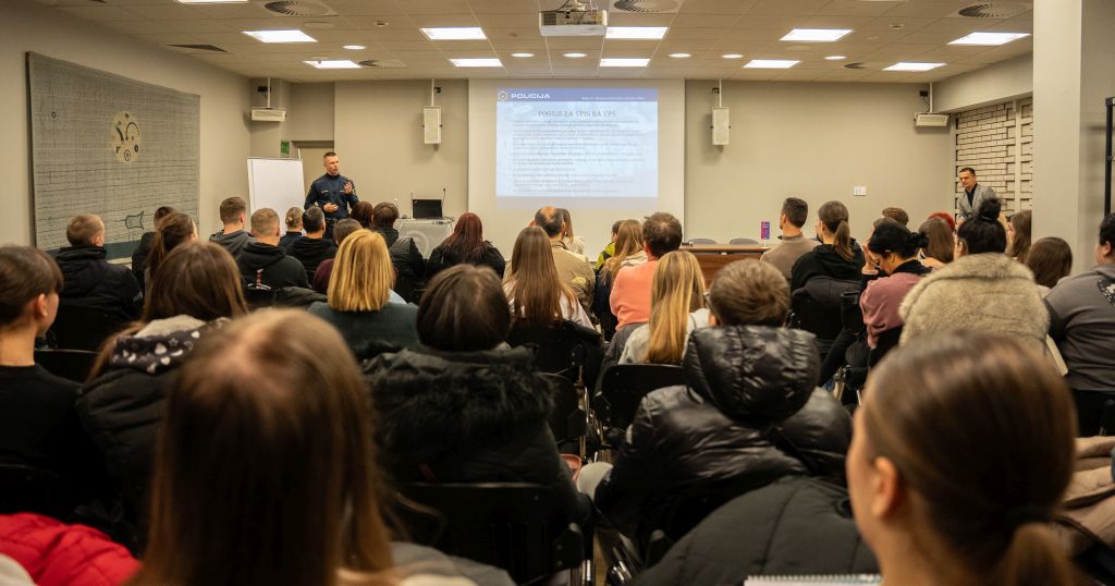 Polna predavalnica poslušalcev, obrnjeni so proti policistu, ki stoji ob projekcijskem platnu in jim govori.