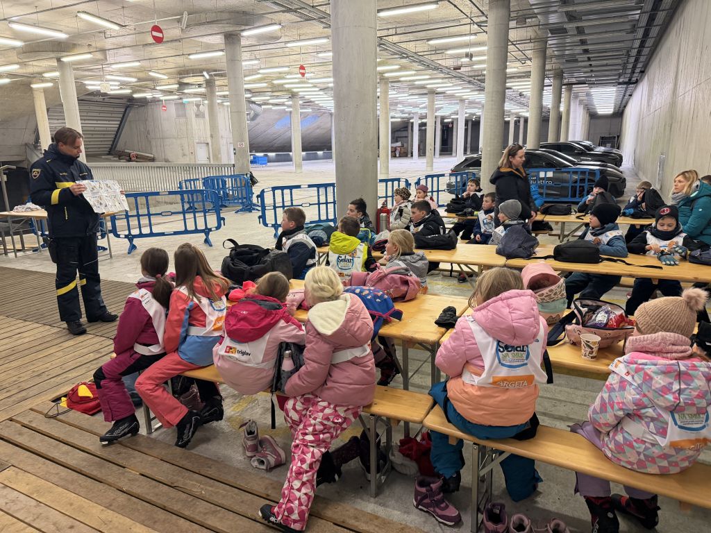 Policist stoji pred skupino otrok v smučarskih kombinezonih, ki sedijo, in jim kaže plakat z napisanimi pravili za vedenje na smučišču.