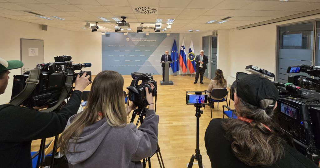 Medijsko središče MNZ med izjavo za javnost; snemalci s kamerami snemajo govorca, ob njem stoji predstavnik policije za odnose z javnostmi.