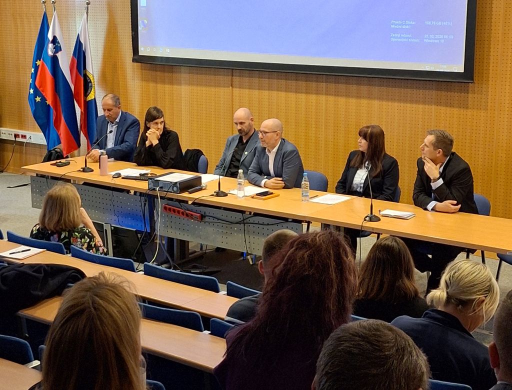 Strokovnjaki na posvetu sedijo za mizo pred polno dvorano poslušalcev