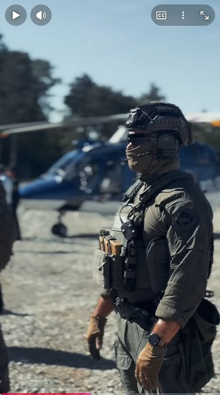 Zajem zaslona: v ospredju stoji policist Specialne enote, v ozadju je policijski helikopter