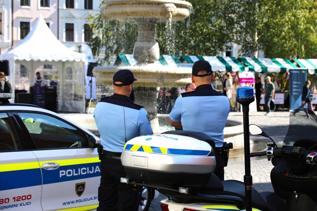 Policista stojita ob policijskem motorju in policijskem avtu, obrnjena sta proti mimoidočim