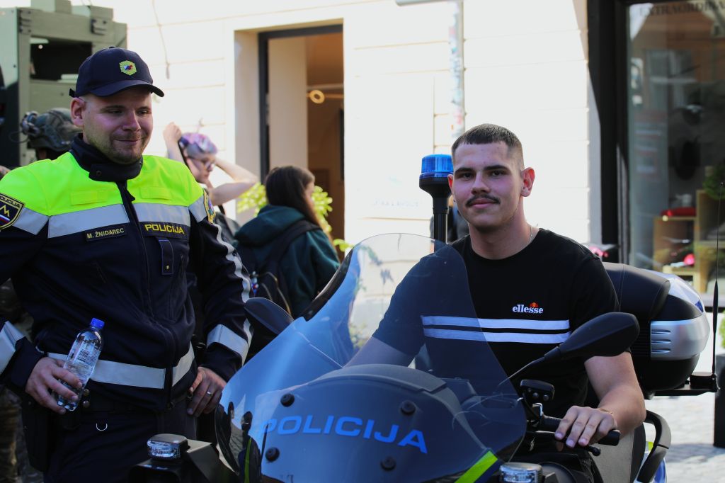 Mladi obiskovalec sedi na policijskem motorju, ob njem stoji policist 