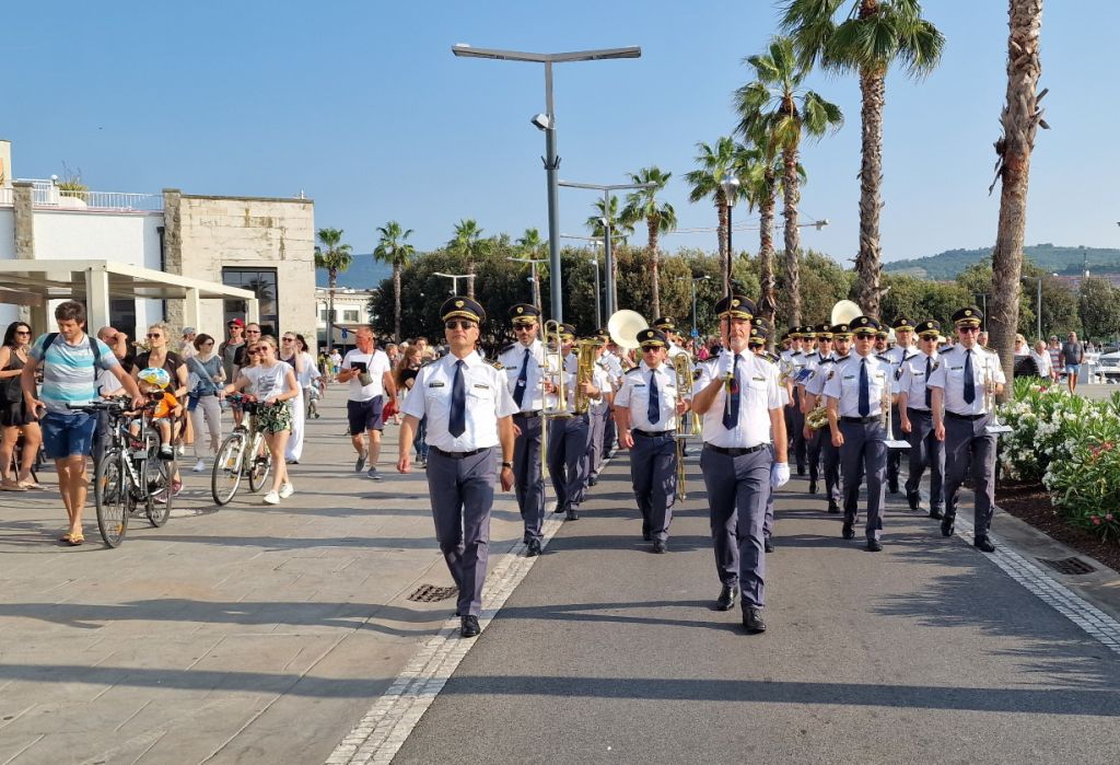 Policijski orkester igra in medtem koraka po koprski obalni promenadi, polni mimoidočih ljudi.