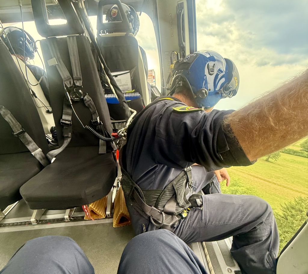 Policist v kabini helikopterja med preletom, nagnjen je iz kabine in opazuje teren pod sabo.