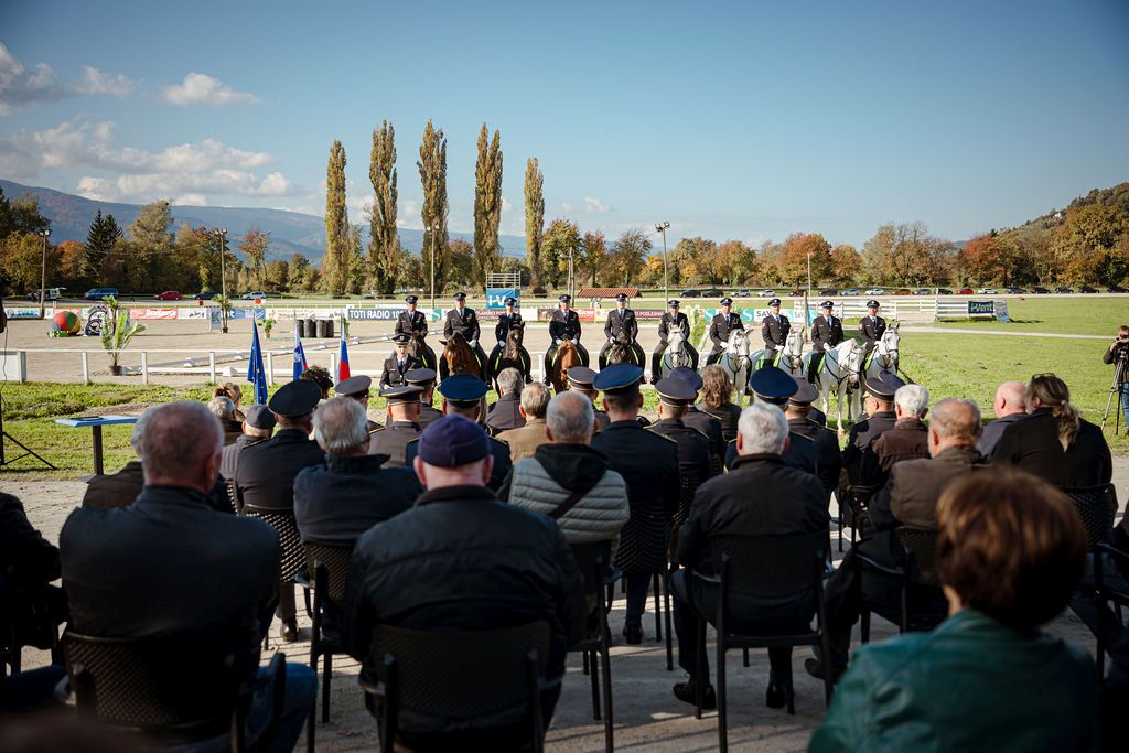 Udeleženci spremljajo proslavo na prostem, v ospredju moderatorka, v ozadju policisti konjeniki