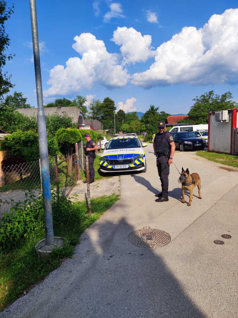 Policist in policist vodnik službenega psa stojita pred ograjo v soseski, v ozadju policijsko vozilo
