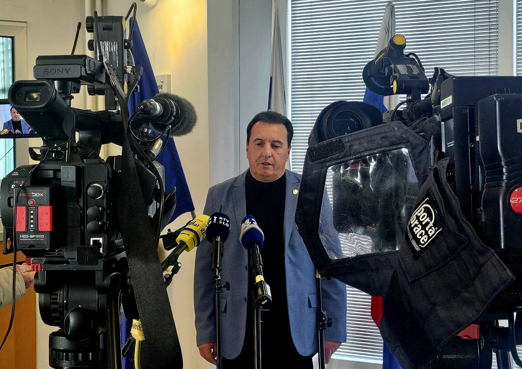 France Božičnik med izjavo za medije, stoji pred kamerami in mikrofoni, v ozadju so zastave.