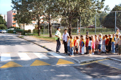 Children accompanied by adults wait in front of a pedestrian crossing for traffic to clear.